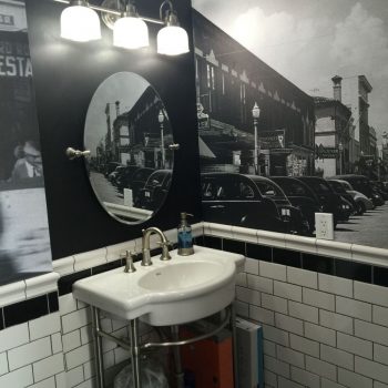 40s bathroom wall wrap featuring a vintage street scene with classic cars, circular mirror above a modern sink, and black and white tiled accents.